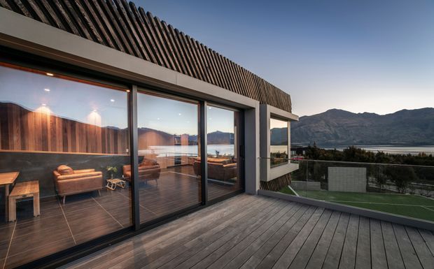 Exterior view of a contemporary multi-level terrace overlooking a lake and mountainous landscape. The broad timber deck, dark-framed floor-to-ceiling glazing, warm timber interior lining, and screened upper roof structure create a strong indoor-outdoor connection.