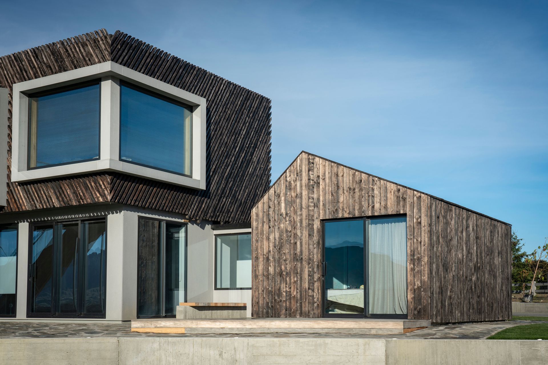 A recessed corner window creates a dramatic aperture in the upper storey façade. Sliding doors in the bedroom wing open out to a deck and overlook the tennis court.