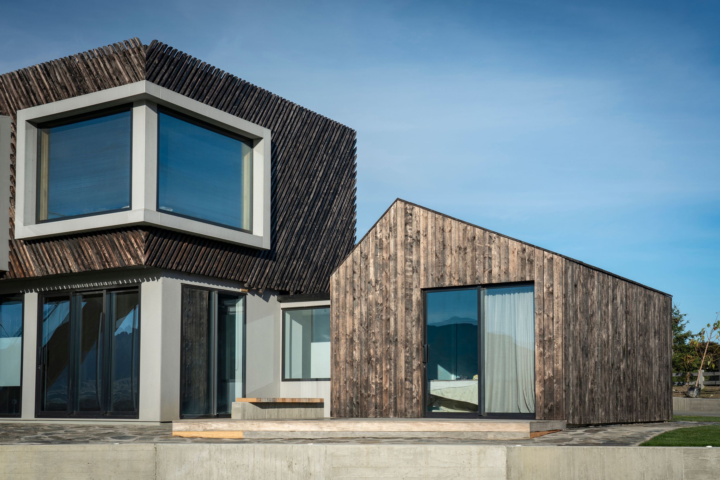 A recessed corner window creates a dramatic aperture in the upper storey façade. Sliding doors in the bedroom wing open out to a deck and overlook the tennis court.