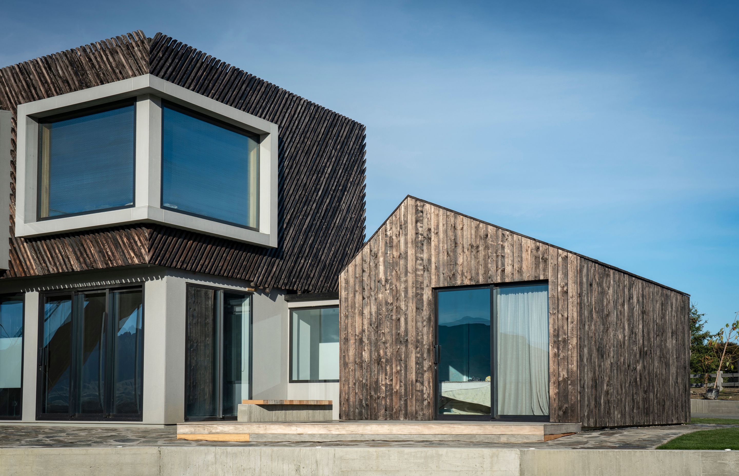 A recessed corner window creates a dramatic aperture in the upper storey façade. Sliding doors in the bedroom wing open out to a deck and overlook the tennis court.