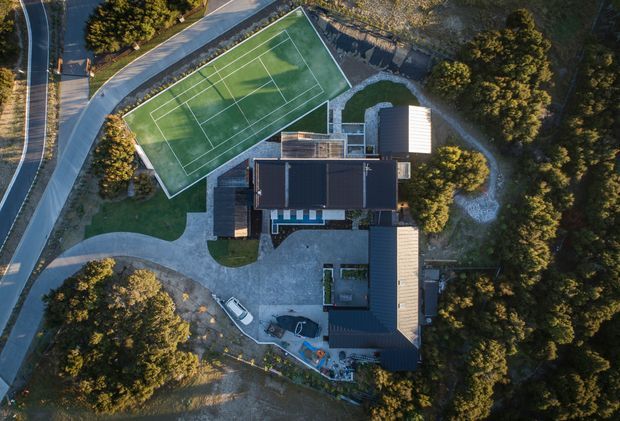 Aerial top-down view of a large contemporary residential property surrounded by dense trees and landscaped grounds. The site includes a prominent fenced multi-court tennis facility, several connected dark-roofed building wings, curved gravel or paved driveways, and landscaped outdoor areas.