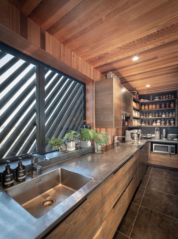A compact, richly detailed kitchen with dark timber cabinetry, extensive timber wall and ceiling lining, open shelving, and a stainless-steel benchtop with an integrated double sink. Diagonal louvred glazing brings in strong natural light, while potted plants, appliances, cookware, and pantry jars add warmth and functionality. The space has a warm, moody, refined character with a strong natural-material palette.