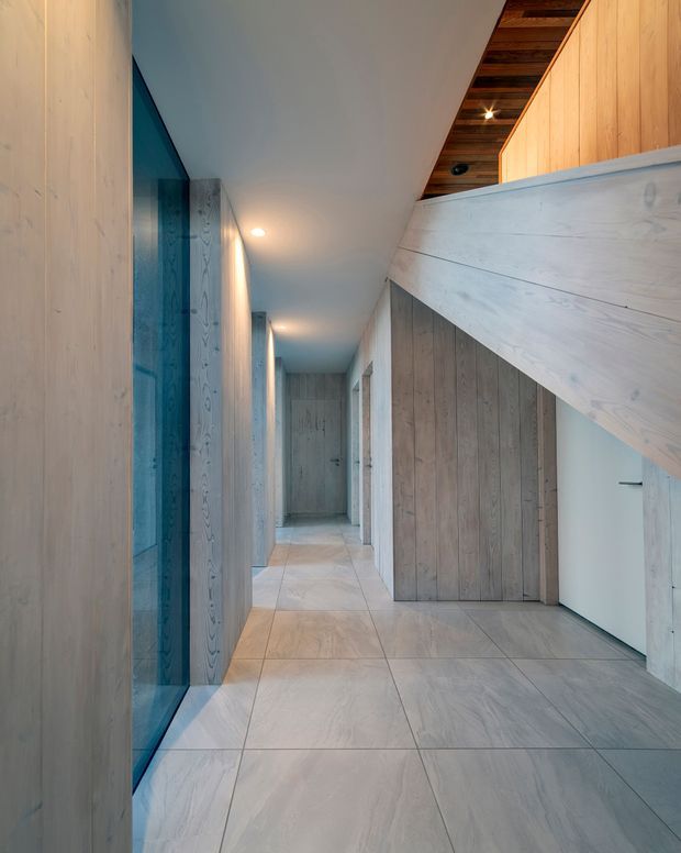 A long, narrow contemporary hallway lined with pale timber wall panelling and large light-coloured tiled flooring. Built-in timber doors and joinery run along both sides, with a blue-tinted glazed opening on the left. A sloping ceiling and exposed dark timber-lined ceiling recess add architectural interest, while recessed downlights provide warm illumination.