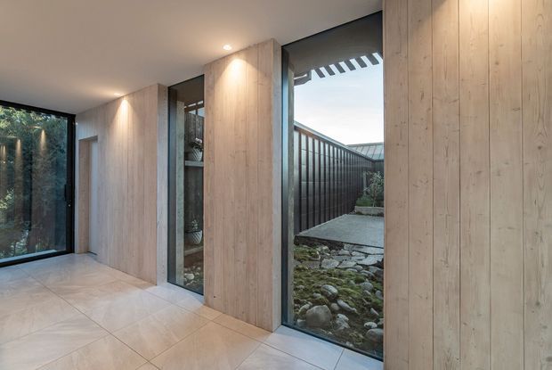 A modern interior hallway or entry passage with pale vertical timber wall panelling, large floor-to-ceiling glazed openings, recessed ceiling lights, and light tiled flooring. The glazing looks onto a narrow landscaped courtyard with dark fencing and stone groundcover. The space has a calm, minimalist material palette and strong indoor-outdoor connection.