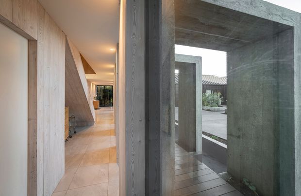 A minimalist architectural hallway with pale timber wall panelling and cabinetry on the left, large light stone or porcelain floor tiles, and a dramatic exposed concrete portal opening toward a small courtyard. Recessed ceiling lighting creates a warm, linear perspective, while the courtyard is visible through the concrete frame.