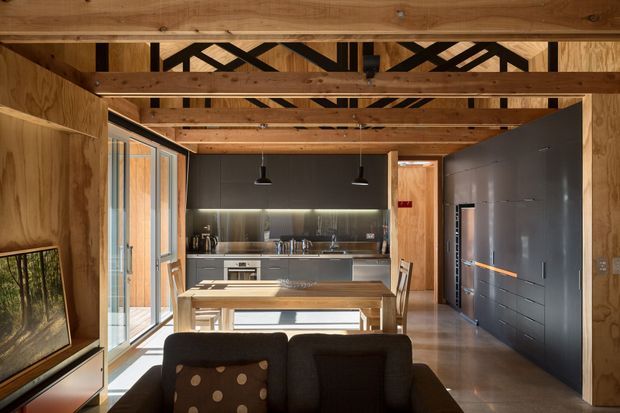 A warm, open-plan interior viewed from the living area toward a modern kitchen and dining zone. Exposed timber beams and structural framing span the ceiling, with timber-lined walls, large glazed sliding doors on the left, a timber dining table, dark cabinetry, stainless-steel appliances, and polished concrete flooring. The space has a refined industrial character with strong natural light and integrated linear lighting.