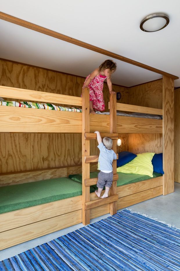A compact children's bedroom featuring custom-built timber bunk beds with integrated ladder and lower storage drawers. The room has warm plywood wall panelling, a white ceiling with timber trim, green bedding, and a blue striped rug over a grey floor. Two children are visible using the bunk ladder.