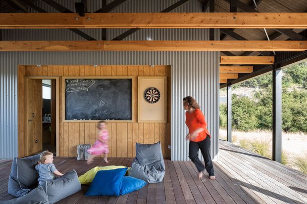A spacious, rustic open-sided timber pavilion or living area with exposed wooden beams, corrugated metal wall panels, vertical timber wall cladding, and a broad timber deck. The room contains a low informal seating area with beanbags and cushions, a chalkboard, and a dartboard, while children and an adult are present. Large open glazing or openings connect the space to the surrounding landscape.
