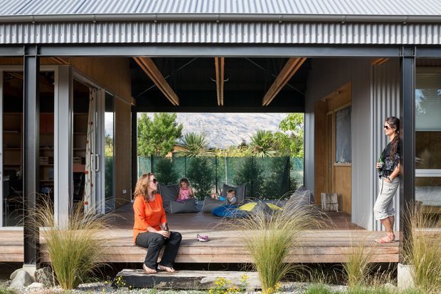 A contemporary corrugated-metal pavilion or extension opening onto a timber deck, with large sliding glazed doors, exposed timber beams, and a relaxed seating area. Several people are present, with ornamental grasses and gravel landscaping in the foreground and a vegetated outlook beyond.