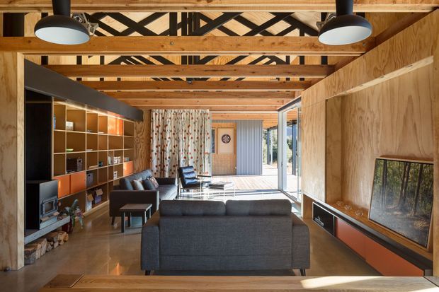 A warm, wood-lined living room with exposed timber roof beams and a distinctive black geometric truss pattern above. The room includes a dark sectional sofa, built-in shelving and cabinetry, a television, lounge chairs, polished concrete flooring, and full-height glazing opening toward an outdoor deck. The interior has a relaxed contemporary character with strong timber detailing and integrated storage.