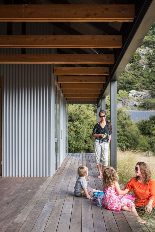 A covered timber deck or verandah runs alongside a corrugated metal-clad building. Exposed timber roof beams and rafters create a strong linear canopy supported by dark vertical posts. Full-height glazing and sliding doors open onto the deck, while several people sit and stand near the edge. Dense bush and rocky terrain are visible beyond the structure, suggesting a home integrated into a natural, sloped setting.