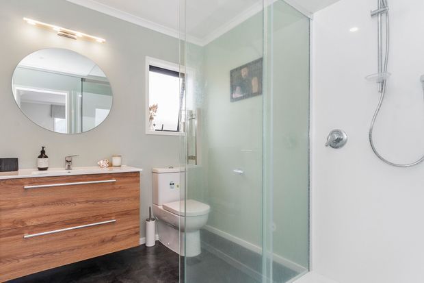 Bright contemporary bathroom with a floating warm timber vanity, round wall mirror, white toilet, and a frameless glass walk-in shower. The room features pale grey walls, dark floor tiles, chrome fixtures, and simple modern lighting.