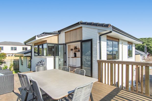 Bright exterior view of a contemporary multi-level home from a raised timber deck. The house has white horizontal cladding, dark window and door frames, a low pitched tiled roof, timber privacy screening, and large glazed openings. A dining table and chairs occupy the foreground deck, with neighbouring suburban homes visible beyond.