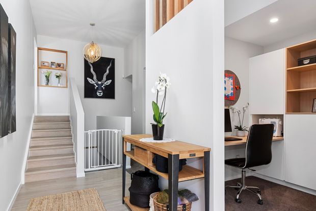 Bright contemporary home entry and stair hall with pale timber flooring, white walls, a timber-and-black console table, decorative pendant lighting, and a carpeted staircase leading upward. A compact home office with built-in timber shelving, desk, and office chair is visible to the right, while a baby gate and decorative artwork sit near the stairs.