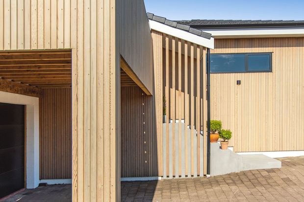 Exterior view of a contemporary residential entry and garage facade, featuring vertical light timber cladding, a dark garage door, a sheltered timber-lined opening, vertical timber screening, dark roof detailing, and a paved forecourt.