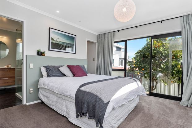 Bright contemporary bedroom with a neatly dressed queen or king bed, layered white bedding, grey and burgundy accent cushions, and a dark grey throw. The room has soft grey walls, carpeted flooring, full-height curtains, a large sliding glazed door opening to an outdoor balcony or terrace, and a spherical pendant light. An adjoining bathroom or dressing area is visible through the open doorway on the left.