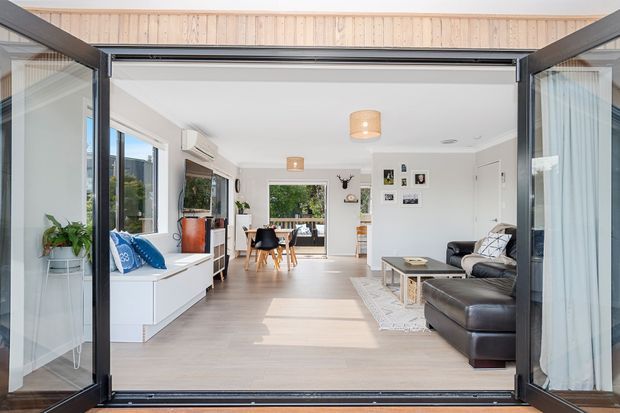 Bright contemporary open-plan living and dining area viewed through fully opened black-framed folding glass doors. The room has pale timber flooring, white walls and built-in cabinetry, a grey sectional sofa, dining table and chairs, simple pendant lights, and strong indoor-outdoor connection to a leafy exterior.