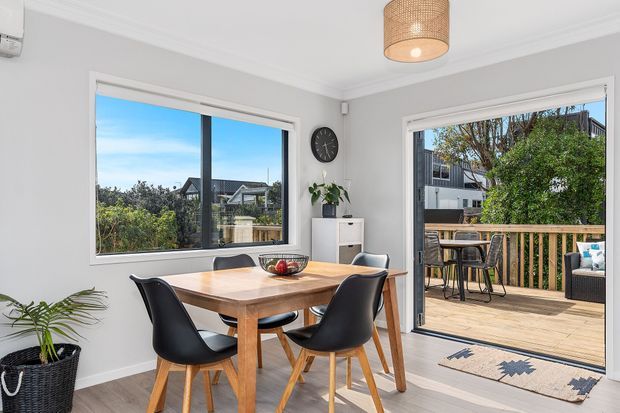 Bright dining area with a timber dining table and four black chairs, opening through wide glazed doors onto a timber deck. The room has light grey walls, white trim, a woven pendant light, indoor plants, and views toward neighbouring homes and greenery.