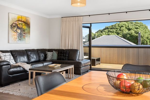 Bright contemporary living and dining area with a large black leather sectional sofa, timber dining table and black dining chairs. Full-height sliding glass doors open to a balcony with a slatted timber privacy screen and views toward a tiled roof and leafy trees. Neutral walls, curtains, a patterned rug, pendant light and artwork complete the space.