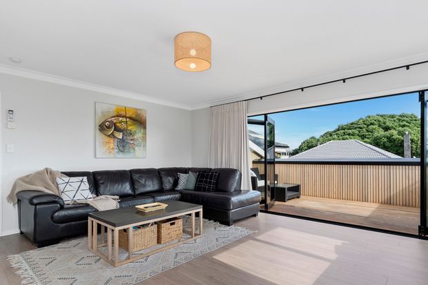 Bright contemporary living room with a large black leather sectional, woven rug, metal-framed coffee table, neutral walls and curtains, and a distinctive cylindrical woven pendant light. Wide sliding glass doors open onto a timber balcony or terrace overlooking a leafy residential setting.