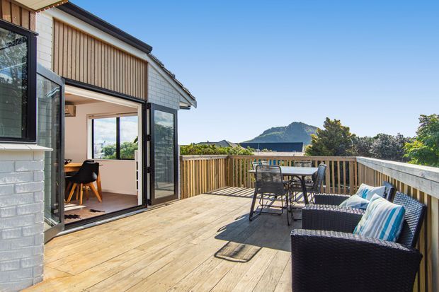 A bright elevated timber deck adjoining a contemporary home, with outdoor dining and wicker lounge seating overlooking a leafy landscape and distinctive mountain peak. Large glazed doors open into an interior dining area, while the exterior shows pale brickwork, vertical timber cladding, and a low-pitched roof.