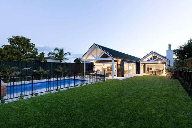 Exterior dusk view of a single-storey suburban home with prominent gable roofs, exposed triangular timber-style truss detailing, dark roof cladding, light walls, and an attached garage or secondary pavilion. A blue swimming pool with black fencing occupies the left side, while a broad manicured lawn and landscaped garden surround the building. Warm interior lighting is visible through the open living areas and garage.