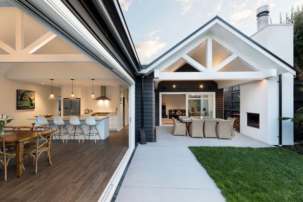 Contemporary residential extension with a light-filled open-plan kitchen and dining area opening directly onto a covered outdoor entertaining space. The home features white walls, dark timber flooring, a pale kitchen island with stools, exposed triangular roof trusses, dark exterior cladding, and an outdoor fireplace beside a paved lawn area.