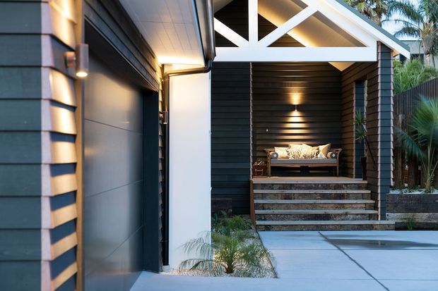 Contemporary covered outdoor entry or lounge area beside a dark-clad garage or building facade. The space features horizontal dark weatherboard cladding, a white timber-framed pitched gable roof structure, integrated warm wall lighting, broad concrete paving, and a raised timber bench with cushions at the rear. Tropical planting borders the paved area.