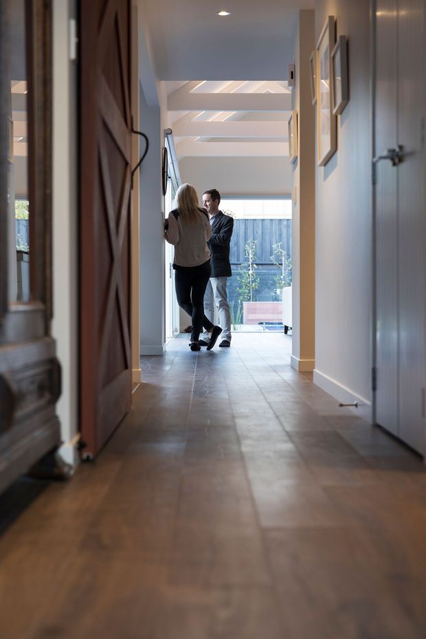 A long residential hallway viewed from near an open timber door, with warm wood flooring, pale walls, framed artwork, and a bright glazed area at the far end. Linear skylights or clerestory glazing illuminate the corridor, where two people are standing near the end.