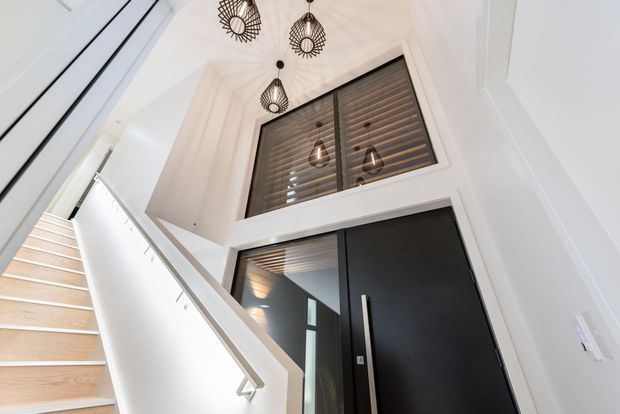 Upward-looking view of a bright contemporary entry stairwell. Pale timber stairs and a white angular balustrade lead to a tall black pivot-style entry door, with black-framed clerestory glazing above featuring horizontal timber screening. Decorative geometric pendant lights hang from the high ceiling, and white wall panelling and trim create a crisp, refined interior.
