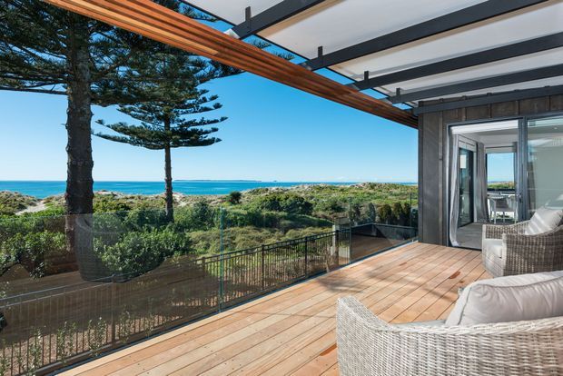 A spacious coastal outdoor terrace overlooking low coastal vegetation and the ocean. The deck features timber flooring, a dark-framed glazed opening into the home, a transparent balustrade, and a striking timber pergola with dark structural beams. Wicker lounge furniture occupies the foreground, creating a relaxed beachside setting.