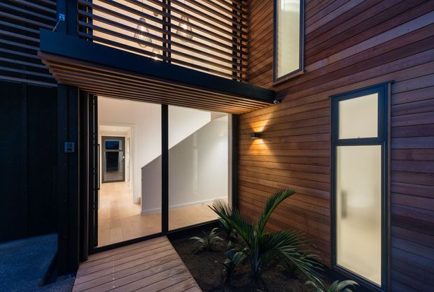 A moody contemporary residential entry courtyard viewed from outside, featuring warm horizontal timber cladding, a timber-decked walkway, black-framed glazed doors, frosted vertical windows, and a slatted overhead balcony or screen. A small planted courtyard sits beside the entry, with a bright hallway visible through the open glass doors.