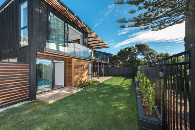A contemporary multi-level residential exterior with dark vertical cladding, warm timber wall sections, extensive glazing, and projecting timber slatted eaves. The landscaped rear yard includes a lawn, planted border, and dark perimeter fencing. Bright coastal-looking daylight and mature trees surround the property.