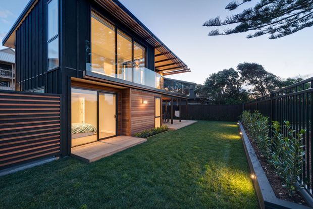 Contemporary two-storey residential extension or townhouse with dark vertical cladding, warm timber-lined soffits and wall accents, extensive glazing, and a compact landscaped rear courtyard. The evening view shows a sheltered lawn, timber deck, screened fencing, planted garden beds, and integrated outdoor lighting.