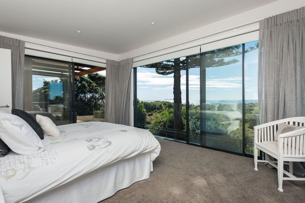 Bright coastal bedroom with a large upholstered bed, white bedding, carpeted floor, full-height black-framed glazing, and grey curtains. The glazing opens to expansive ocean and landscaped views, while a white timber bench sits beside the windows.