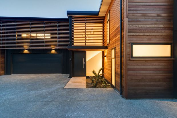 Contemporary residential exterior viewed from the driveway, featuring dark stained horizontal timber cladding, extensive horizontal louvre screens, black-framed glazing and garage doors, and a recessed illuminated entry with a small planted area. The composition has strong rectilinear volumes and warm evening lighting.