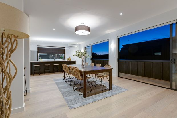 Open-plan dining and kitchen interior photographed at dusk. A timber dining table with woven chairs sits on a large rug, with a compact kitchen and island seating behind it. Full-height glazed doors and windows open to a dark exterior, while recessed ceiling lights and pendant fixtures provide warm illumination. The room features pale timber flooring, white walls and cabinetry, and dark window and door framing.
