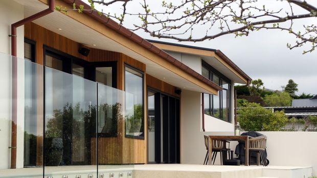 Contemporary residential exterior showing a covered terrace or balcony with frameless glass balustrading, timber-clad walls, dark-framed glazing, and a low-pitched tiled roof. Outdoor dining furniture sits against a pale rendered wall, with established planting visible around the house.
