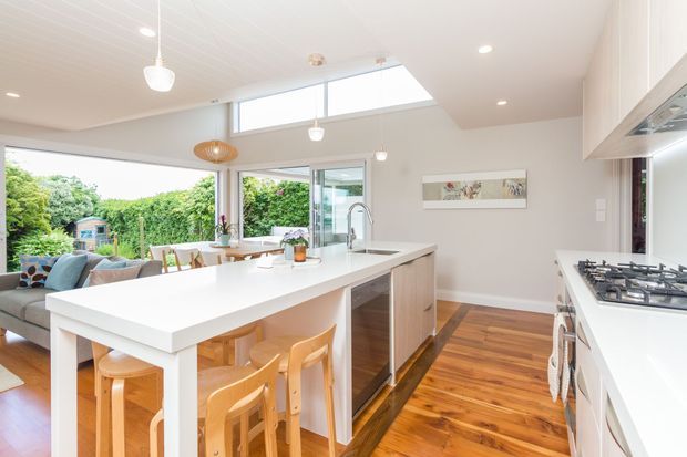 Bright open-plan kitchen with a large white island and integrated sink, timber bar stools, white cabinetry, warm timber flooring, and a gas cooktop along the right wall. The space opens to a dining and living area with extensive glazing, a skylight, and views of a leafy garden.