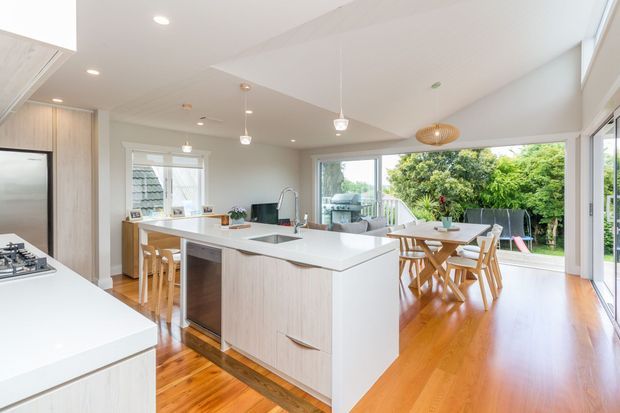 Bright open-plan kitchen and dining area with a large white island, integrated stainless-steel appliances, pale timber cabinetry, warm timber flooring, pendant lighting, and extensive glazing opening to a leafy outdoor area. The space has a clean contemporary feel with abundant natural light.
