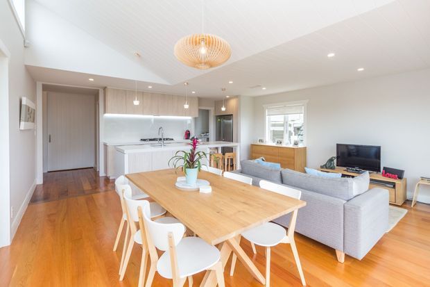 Bright open-plan interior combining a kitchen, dining area and living room. The space features warm timber flooring, white walls and ceiling, pale fixed cabinetry, a large timber dining table with white chairs, and a light grey sectional sofa. Recessed lighting and a sculptural pendant add contemporary detailing, while a window brings in natural light.