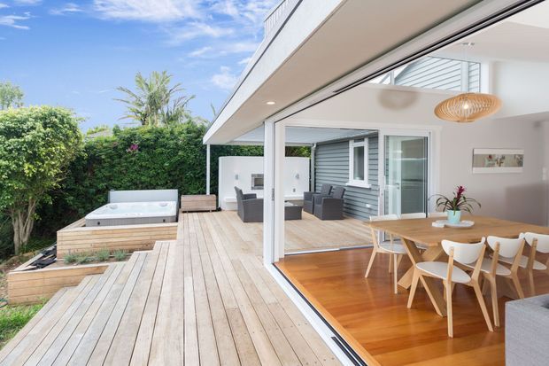 A bright contemporary indoor-outdoor living and dining area opening onto a large timber deck. The space features full-height sliding glass doors, timber flooring, a light timber dining table with sculptural chairs, a woven pendant light, and a built-in outdoor spa beside landscaped greenery. Grey horizontal cladding is visible on the adjoining exterior wall.
