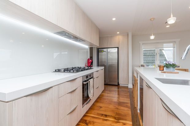 Bright contemporary kitchen with pale timber-look cabinetry, white benchtops and splashback, stainless-steel appliances, a central island with integrated sink, pendant lighting, and warm timber flooring. A window above the sink brings in natural light.