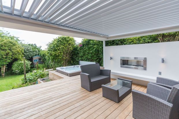 Covered outdoor entertaining area with a broad timber deck, grey woven outdoor lounge furniture, built-in wall-mounted barbecue, and a raised spa beside landscaped greenery. The space is sheltered by a modern white-framed louvred roof and opens onto a garden.