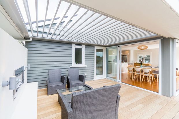 Bright contemporary outdoor entertaining area beneath a white louvred pergola, with timber decking, grey weatherboard cladding, and woven outdoor lounge furniture. Large glazed sliding doors open into a warm dining area with timber flooring and a pale timber dining table and chairs.