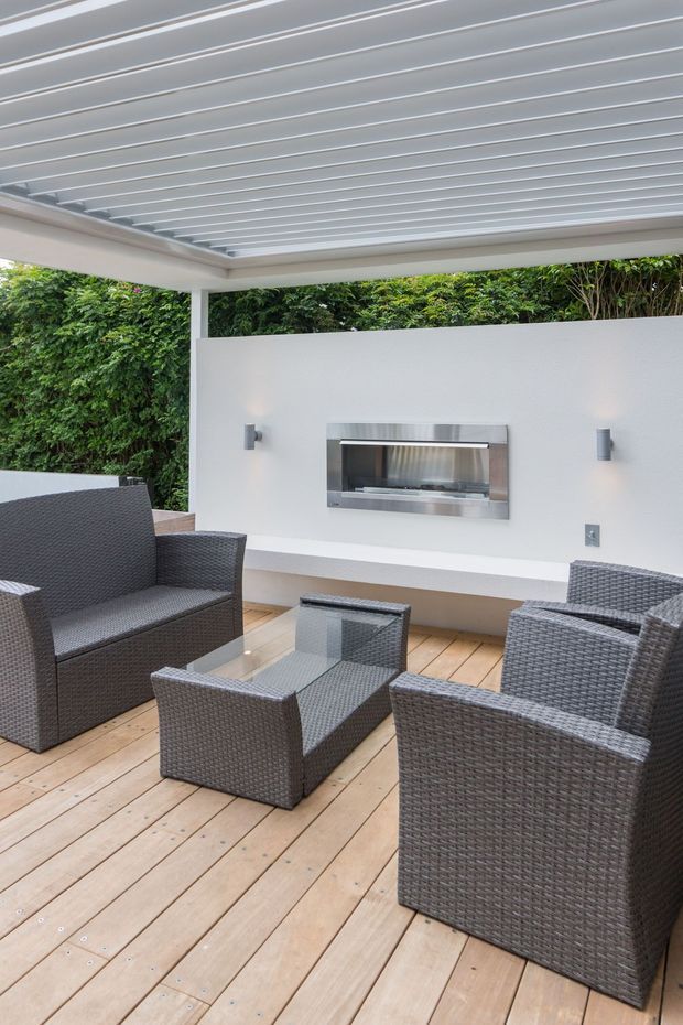 A covered outdoor entertaining area with a timber deck, dark woven outdoor lounge chairs and a coffee table, plus a built-in stainless steel barbecue set into a white rendered wall. The space has a white louvred roof, integrated lighting, and dense green screening behind it.