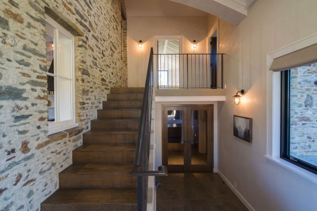 A warm, rustic entry hall with a timber staircase rising beside an exposed natural stone wall. The space includes dark metal balustrades, a raised landing, timber-framed glazed doors, wall sconces, and windows overlooking the stone exterior wall.