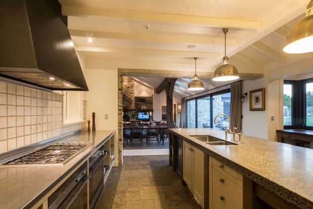 A spacious traditional kitchen with a large central island, extensive stone benchtops, integrated gas cooktop, tiled splashback, pendant lighting and exposed ceiling beams. The kitchen opens to a dining and living area with extensive glazing and a stone-clad feature wall.