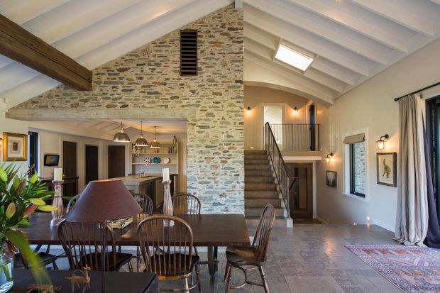 Open-plan rustic interior showing a dining area, kitchen, and double-height circulation space. The room features exposed rough stone masonry, white painted exposed ceiling beams, a substantial timber beam, a pitched ceiling, timber dining furniture, pendant lighting, and a staircase with dark metal balustrades. A skylight and tall windows bring in natural light, while a patterned rug and curtains add warmth.