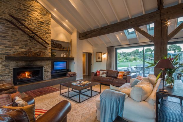 Warm, rustic living room with a large stacked-stone fireplace, timber mantel, exposed dark timber beams, white paneled ceiling, and expansive glazing opening to a landscaped garden. Leather armchairs, sofas, a patterned area rug, coffee table, and television create a comfortable, inviting lounge setting.
